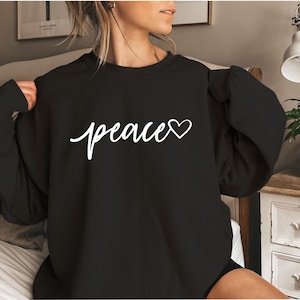May include: Black crew neck sweatshirt with the word "peace" in white script and a white heart symbol. The sweatshirt is a solid black color and has long sleeves. The text is centered on the front of the garment.