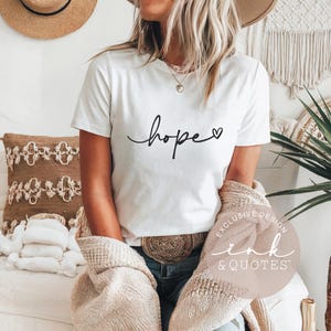May include: White t-shirt with the word "hope" in a cursive font and a small heart. The shirt is part of a clothing collection. The image also includes a decorative pillow, a hat, and a beige sweater.