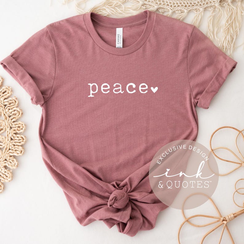 May include: A dusty rose t-shirt with the word "peace" in white script, a small white heart follows the word. The shirt is knotted at the bottom. The words "EXCLUSIVE DESIGN ink & QUOTES" are in a circular design on the bottom right.
