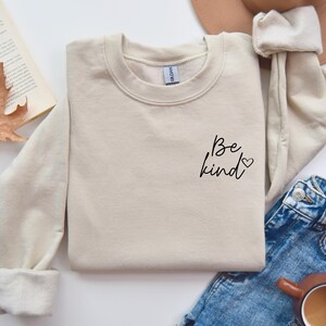 May include: A beige sweatshirt with the text "Be kind" and a small heart on the left side of the chest.