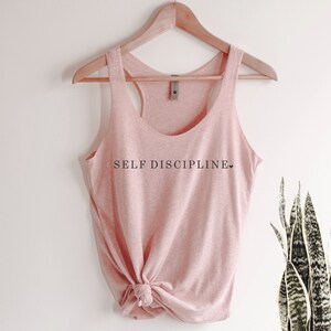 May include: A pink tank top with the text "SELF DISCIPLINE" printed on the front. The tank top is hanging on a wooden hanger.