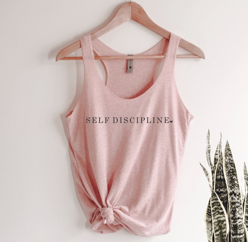 May include: A pink tank top with the text "SELF DISCIPLINE" printed on the front. The tank top is hanging on a wooden hanger.