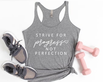 May include: Gray tank top with the text "STRIVE FOR progress NOT PERFECTION" in white with a small pink heart. The tank top is laying on a white surface with a pair of gray running shoes and pink dumbbells.