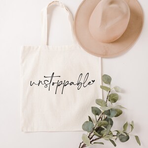Op de afbeelding: Een witte canvas tote bag met zwarte tekst die "unstoppable" zegt met een klein hartje. De tas ligt plat op een wit oppervlak met een bruine hoed en een takje groene bladeren.