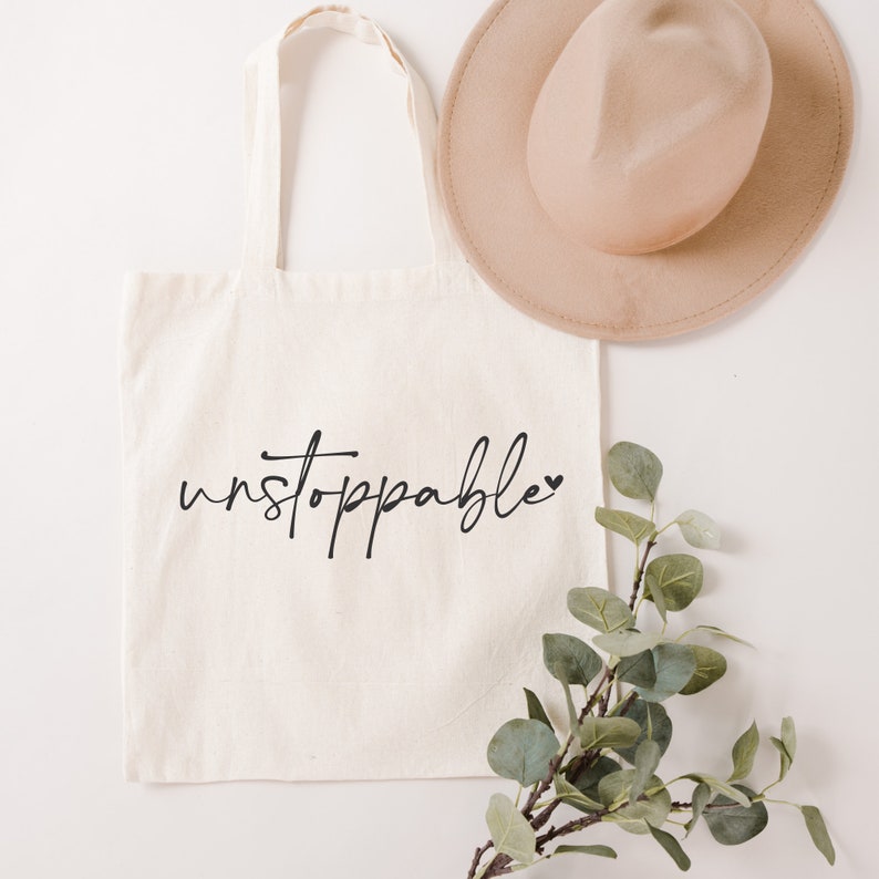 Op de afbeelding: Een witte canvas tote bag met zwarte tekst die "unstoppable" zegt met een klein hartje. De tas ligt plat op een wit oppervlak met een bruine hoed en een takje groene bladeren.
