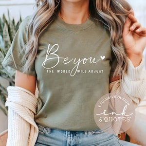 May include: Olive green t-shirt with the white text "Be you" and "The world will adjust". The shirt has short sleeves and a crew neck. The design includes a small heart. The shirt is part of the "Ink & Quotes" exclusive design collection.
