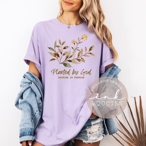 May include: A light purple t-shirt with the text "Planted by God" and a floral design in shades of brown and yellow. The shirt also has the text "Growing in Purpose".