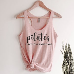 K&ouml;nnte beinhalten: Ein hellrosa Tanktop mit dem Text "pilates is my love language" in schwarzer Schrift. Das Tanktop h&auml;ngt an einem Holzkleiderb&uuml;gel. Der untere Teil des Tanktops ist geknotet.