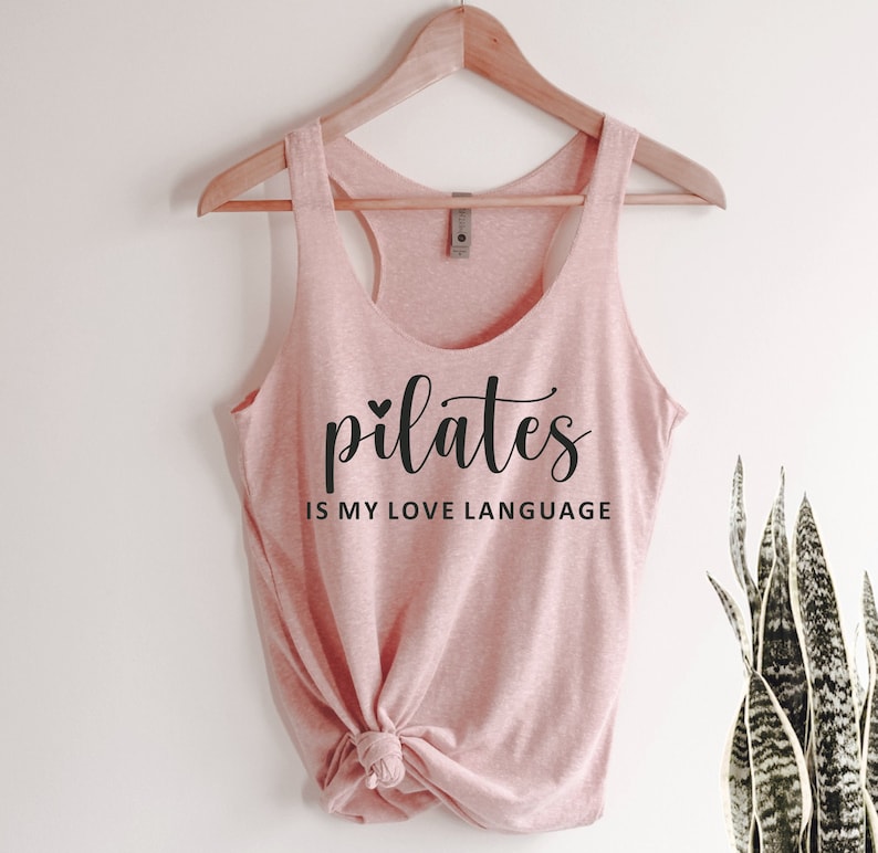 K&ouml;nnte beinhalten: Ein hellrosa Tanktop mit dem Text "pilates is my love language" in schwarzer Schrift. Das Tanktop h&auml;ngt an einem Holzkleiderb&uuml;gel. Der untere Teil des Tanktops ist geknotet.