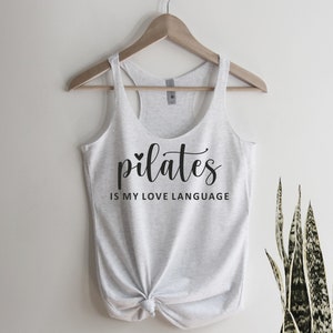 K&ouml;nnte beinhalten: Ein hellgraues Tanktop mit der Aufschrift "pilates IS MY LOVE LANGUAGE" in schwarzer Schrift. Das Tanktop ist am unteren Ende geknotet und h&auml;ngt an einem Holzb&uuml;gel. Im Hintergrund ist eine gr&uuml;ne Pflanze zu sehen.