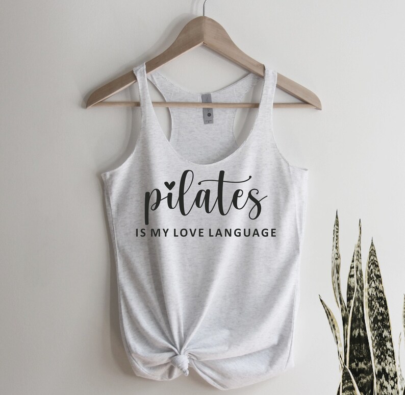 K&ouml;nnte beinhalten: Ein hellgraues Tanktop mit der Aufschrift "pilates IS MY LOVE LANGUAGE" in schwarzer Schrift. Das Tanktop ist am unteren Ende geknotet und h&auml;ngt an einem Holzb&uuml;gel. Im Hintergrund ist eine gr&uuml;ne Pflanze zu sehen.