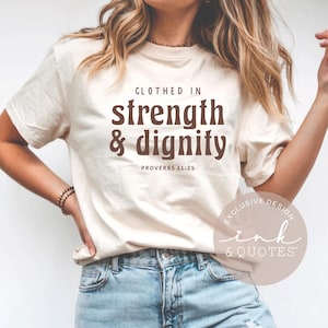 Pode incluir: Camiseta creme com o texto "Clothed in strength & dignity" em letras marrons, com "Proverbs 31:25" abaixo. A camisa é combinada com jeans azuis. O design inclui o logotipo "ink & QUOTES".