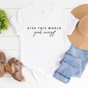 May include: White t-shirt with the text "GIVE THIS WORLD good energy" in black lettering with a small heart symbol.