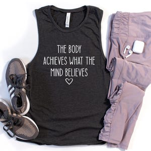 May include: A dark gray tank top with the text "The body achieves what the mind believes" and a small heart symbol. The tank top is paired with a pair of light purple leggings.