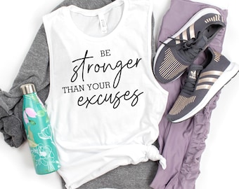 May include: White tank top with the text "Be stronger than your excuses" in black. The tank top is laying on a gray and white shirt. Next to the tank top are purple leggings and a pair of black and gold running shoes.