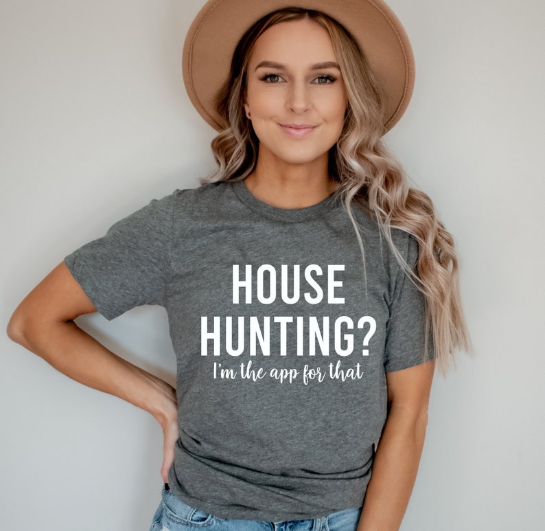 May include: A woman wearing a gray t-shirt with the text "HOUSE HUNTING? I'm the app for that" in white. She is smiling and has her hands on her hips.
