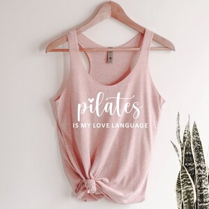 K&ouml;nnte beinhalten: Hellrosa Tanktop mit wei&szlig;em Schriftzug "pilates IS MY LOVE LANGUAGE". Das Tanktop h&auml;ngt an einem Holzkleiderb&uuml;gel. Im Hintergrund ist eine Pflanze zu sehen.