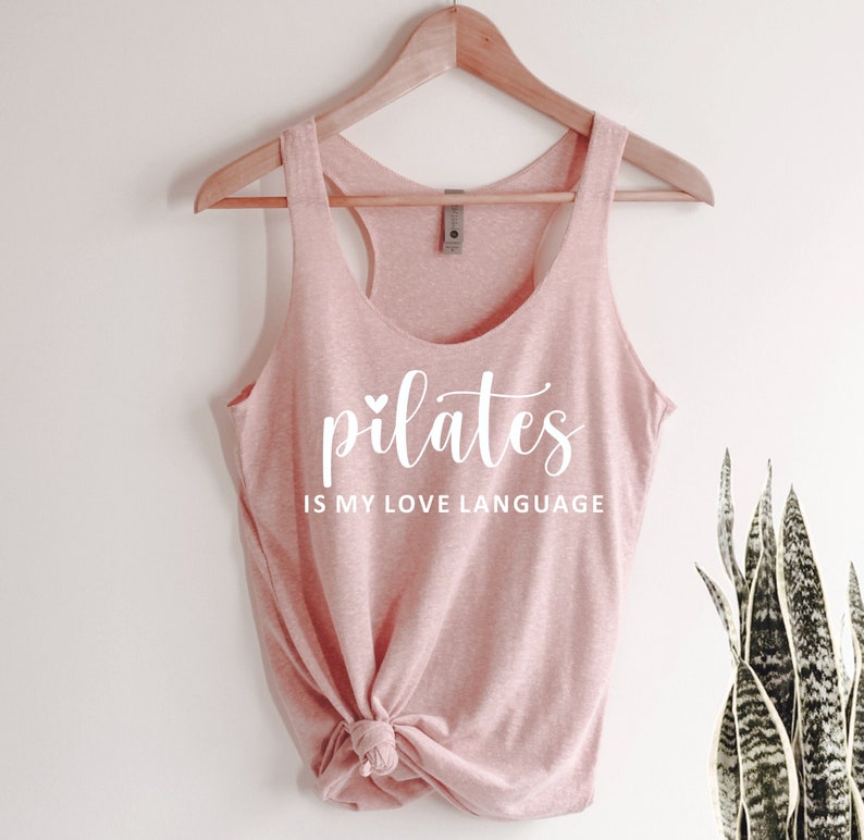 K&ouml;nnte beinhalten: Hellrosa Tanktop mit wei&szlig;em Schriftzug "pilates IS MY LOVE LANGUAGE". Das Tanktop h&auml;ngt an einem Holzkleiderb&uuml;gel. Im Hintergrund ist eine Pflanze zu sehen.