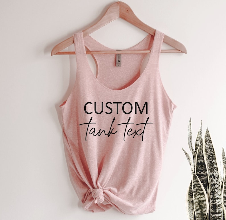 May include: A light pink tank top with the text "CUSTOM tank text" in black script. The tank top is hanging on a wooden hanger. The bottom of the tank top is tied in a knot. A green plant is visible on the right side.
