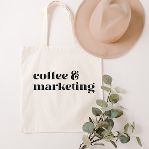 Op de afbeelding: Een witte canvas tote bag met zwarte tekst die "coffee & marketing" zegt. De tas ligt plat op een wit oppervlak met een bruine hoed en een takje groene bladeren.