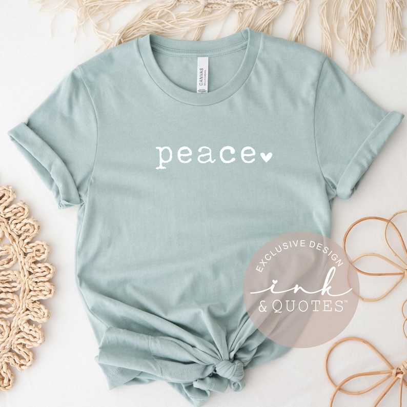 May include: A light blue t-shirt with the word "peace" in white script and a small white heart. The shirt has rolled-up sleeves and a knot at the bottom. The text "EXCLUSIVE DESIGN ink & QUOTES" is printed on a circular sticker.