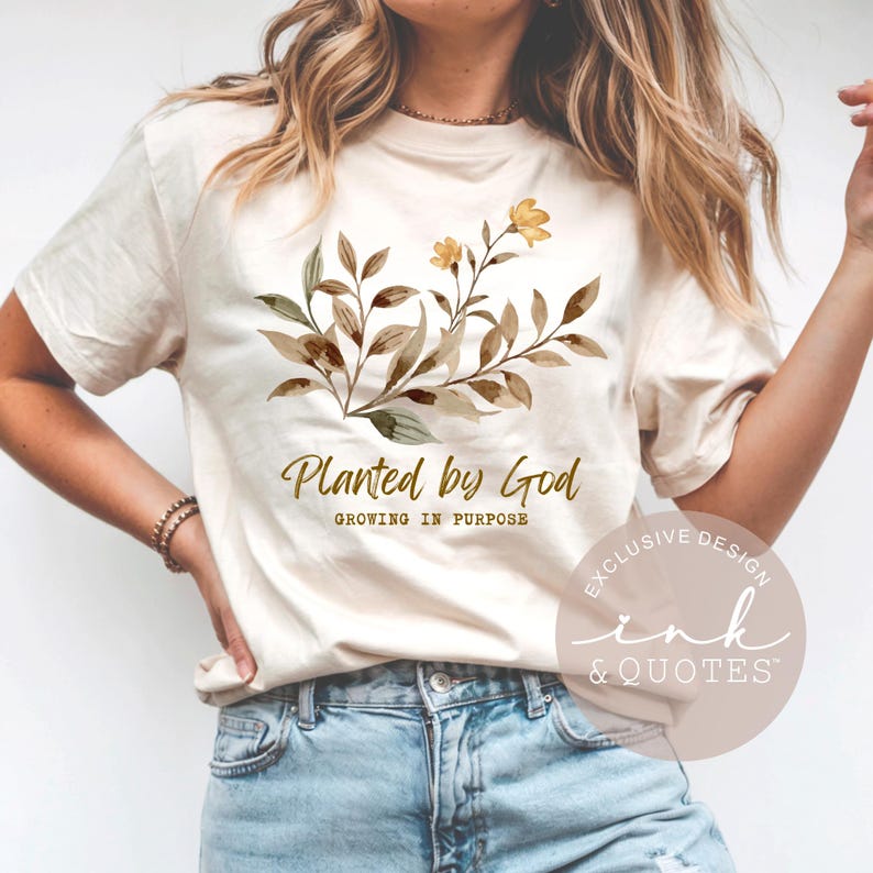May include: Cream-colored t-shirt featuring a watercolor floral design in shades of brown and green, with a yellow flower accent. The text "Planted by God" and "Growing in Purpose" are printed below the floral design.