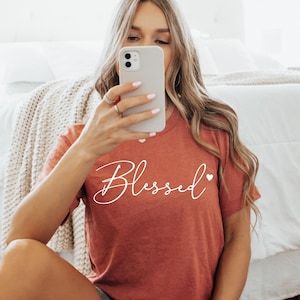 May include: A rust-colored t-shirt with the word "Blessed" written in white cursive script with a small white heart to the right of the word.