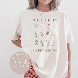 May include: Cream-colored t-shirt featuring the text "AUTHENTICITY IS A REVOLUTION" in brown, with a floral design. The design includes various watercolor flowers in shades of blue, pink, red, and yellow. The shirt also has a circular logo that says "EXCLUSIVE DESIGN ink & QUOTES".