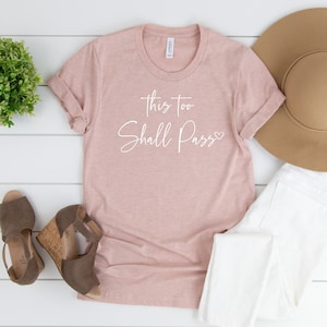 May include: A light pink t-shirt with rolled-up sleeves, featuring the text "this too Shall Pass" in white script. The shirt is paired with white jeans, brown wedge sandals, and a wide-brimmed hat. A small green plant is also visible.
