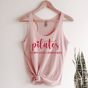 K&ouml;nnte beinhalten: Ein hellrosa Tanktop mit dem roten Schriftzug "pilates is my love language". Das Tanktop ist am unteren Ende geknotet und h&auml;ngt an einem Holzkleiderb&uuml;gel.