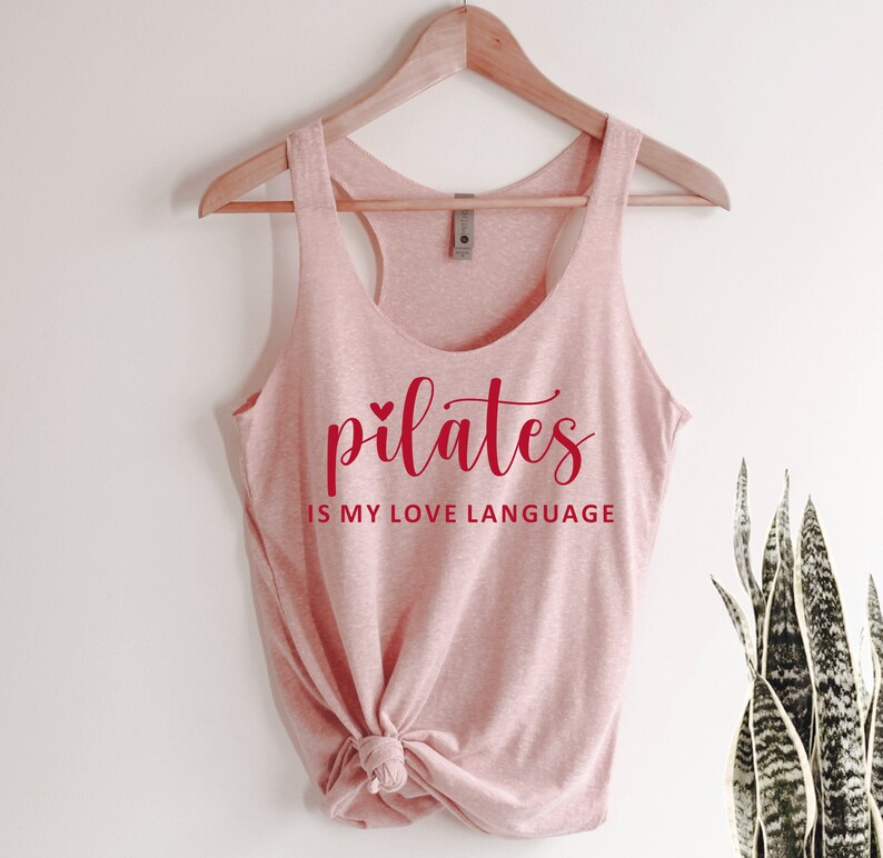 K&ouml;nnte beinhalten: Ein hellrosa Tanktop mit dem roten Schriftzug "pilates is my love language". Das Tanktop ist am unteren Ende geknotet und h&auml;ngt an einem Holzkleiderb&uuml;gel.
