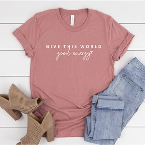 May include: A dusty rose t-shirt with the text "GIVE THIS WORLD good energy" in white lettering with a small heart symbol.