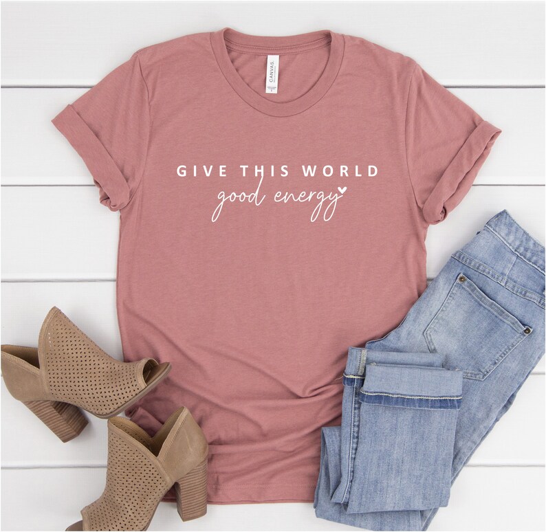 May include: A dusty rose t-shirt with the text "GIVE THIS WORLD good energy" in white lettering with a small heart symbol.