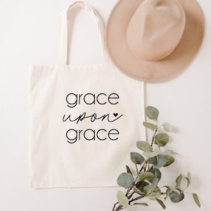 May include: A white canvas tote bag with black text that reads "grace upon grace" with a small black heart symbol. The bag is lying flat on a white surface with a brown hat and a sprig of green leaves.