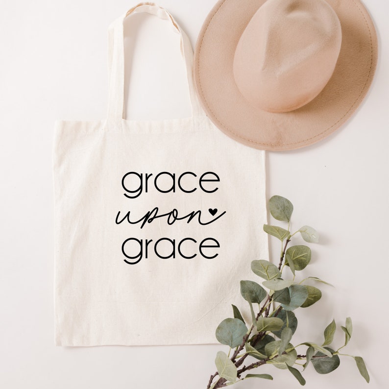 May include: A white canvas tote bag with black text that reads "grace upon grace" with a small black heart symbol. The bag is lying flat on a white surface with a brown hat and a sprig of green leaves.