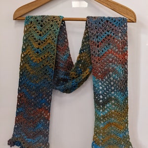 May include: A crocheted scarf with a multi-colored ombre pattern in shades of blue, green, brown, and orange. The scarf has a fringed edge and is hanging on a wooden hanger.