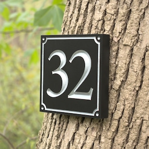 May include: Black square house number sign with the number 32 in silver. The sign is mounted on a tree trunk. The corners of the sign have decorative detailing.
