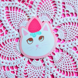 May include: A die-cut sticker featuring a white cat face with brown eyes, pink ears, and a strawberry on its head. The sticker has a pink border and is set against a white crocheted doily on a bright pink background.