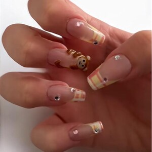 May include: Long, square-shaped nails with a light pink base, decorated with a 3D monkey and small rhinestones. Some nails have a yellow and orange striped pattern. The manicure is detailed and eye-catching.