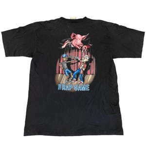Puede incluir: Camiseta negra con un gráfico de un cerdo titiritero controlando dos figuras de dibujos animados en un escenario. El texto "The Rap Game" está debajo. La camiseta tiene un estilo vintage.