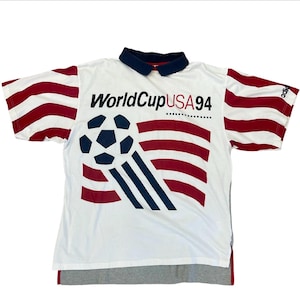 May include: White polo shirt with red and white striped sleeves and a navy blue collar. The shirt features a graphic of a soccer ball and the text "World Cup USA 94" in black and red.