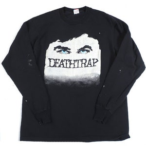 Peut inclure: T-shirt à manches longues noir avec un motif blanc d'un œil et le texte "DEATHTRAP" imprimé en dessous.