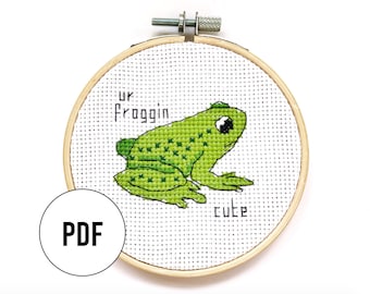 May include: A cross-stitch pattern featuring a green frog with the text "ur froggin cute" on a white background. The pattern is in a wooden embroidery hoop.