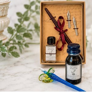 May include: A calligraphy set in a rectangular box containing a dark brown wooden pen, two silver metal nibs, and a small bottle of Fine Italian Writing Ink. A separate blue glass dip pen rests on a glass stand next to a larger ink bottle.