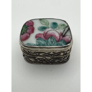 May include: A small, rectangular silver box with a floral design on the lid. The lid features a white background with pink and red roses, green leaves, and a black stem. The sides of the box have an intricate, openwork design.