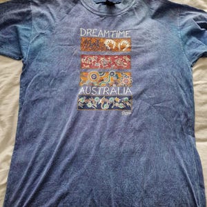 May include: Blue t-shirt with the text "DREAMTIME AUSTRALIA" and colorful Aboriginal art designs. The shirt has a tie-dye effect and a crew neck.