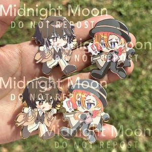 May include: Four enamel pins featuring anime-style characters. Two characters have brown hair and are wearing tan coats. The other two characters have orange hair and are wearing black hats and coats. The pins have gold-colored borders. The text "Midnight Moon DO NOT REPOST" is printed on the pins.