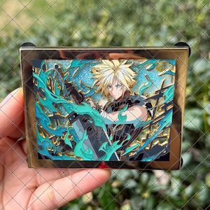 May include: A rectangular metal enamel pin featuring a blonde character with spiky hair holding a large sword, surrounded by swirling teal energy effects against a glittery blue background, framed in a polished gold-tone metal border.