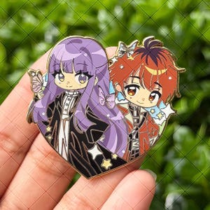 May include: A heart-shaped enamel pin featuring two anime-style characters. One character has long purple hair and a black coat, while the other has short red hair and a red outfit. The design includes small butterfly and star accents with gold metal outlines.