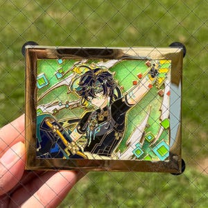 Pode incluir: Um broche de esmalte retangular com moldura dourada apresentando um personagem estilo anime com cabelo escuro e marcas verdes. O personagem veste uma roupa preta com detalhes dourados sobre um fundo verde brilhante com formas geométricas quadradas.